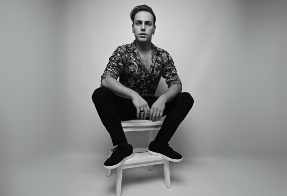 Person in gemustertem Hemd und schwarzer Hose sitzt auf einem Schwarz-Weiß-Foto selbstbewusst auf einem Hocker.